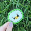 A hand holding a round sticker with an egg character illustration on it, placed against a grassy background.