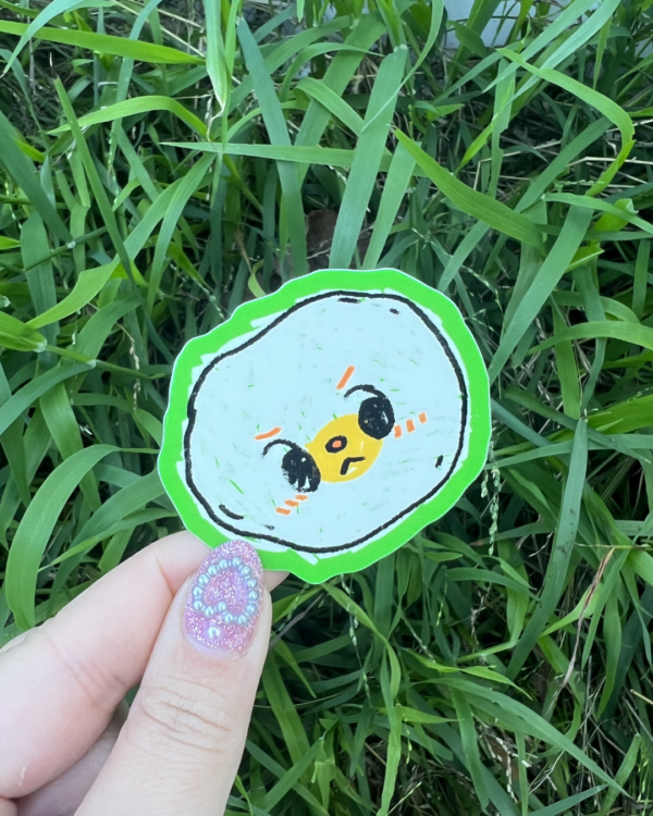 A hand holding a round sticker with an egg character illustration on it, placed against a grassy background.
