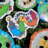 multi colored worm wearing a party hat with "Yippee" and confetti on top sticker laying on grass with other stickers
