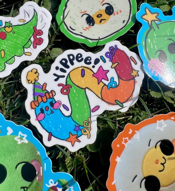 multi colored worm wearing a party hat with "Yippee" and confetti on top sticker laying on grass with other stickers