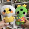 IMG_8067 One hand on the left holding a playful egg plushie in a yellow egg mini cardigan. A right hand holding a playful alien plush with a mismatched rainbow mini cardigan. In the background are two bookshelves with miscellaneous anime figures and trinkets.