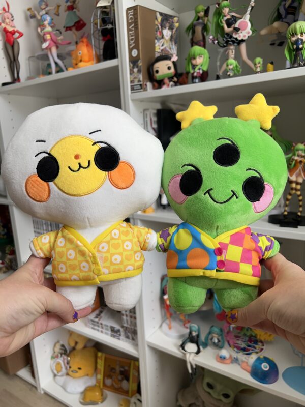 IMG_8067 One hand on the left holding a playful egg plushie in a yellow egg mini cardigan. A right hand holding a playful alien plush with a mismatched rainbow mini cardigan. In the background are two bookshelves with miscellaneous anime figures and trinkets.