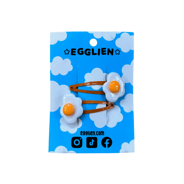 A pair of fried egg cabochons on orange hairclips both on a blue backing card that reads "Egglien"