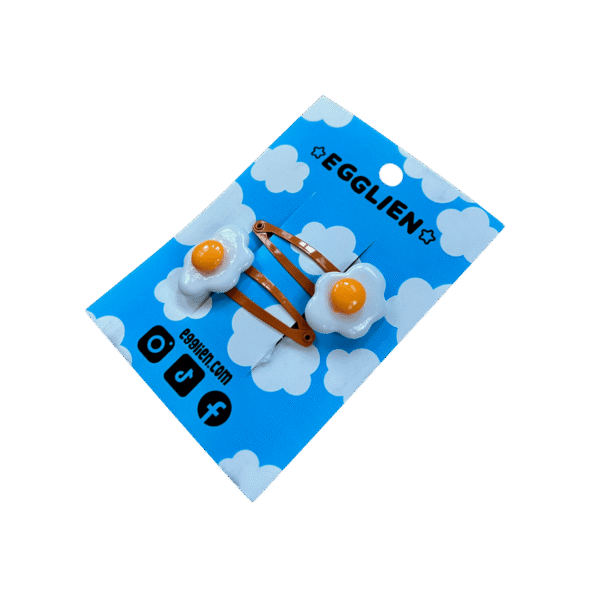 A pair of fried egg cabochons on orange hairclips both on a blue backing card that reads "Egglien"