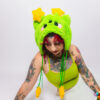 Woman with tattoos sitting on her side with one arm on the ground and head facing the ground to show the plush alien hat with cat ears on her head. She is wearing a matching plush green hat with cat ears, a green tank top, white skirt with a green petti coat poking out.