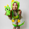 Woman with tattoos playfully with her tongue out and eyes closed toward the camera holding a cheerful alien plush. She is wearing a matching plush green hat with cat ears, a green tank top, white skirt with a green petti coat poking out and white shoes.