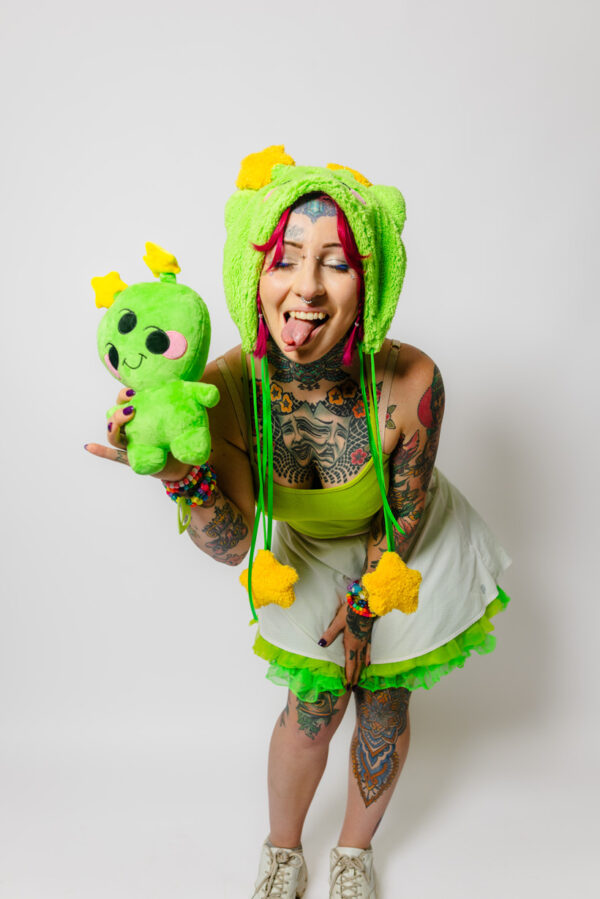 Woman with tattoos playfully with her tongue out and eyes closed toward the camera holding a cheerful alien plush. She is wearing a matching plush green hat with cat ears, a green tank top, white skirt with a green petti coat poking out and white shoes.