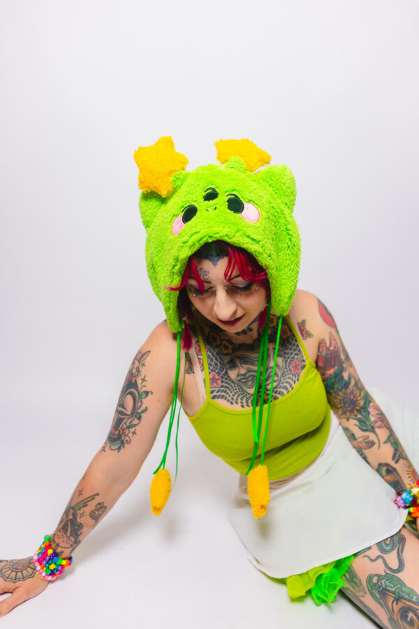 Woman with tattoos sitting on her side with one arm on the ground and head facing the ground to show the plush alien hat with cat ears on her head. She is wearing a matching plush green hat with cat ears, a green tank top, white skirt with a green petti coat poking out.