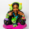 Girl sitting cross legged playfully looking at the camera. She is wearing bright green alien earmuffs, a roller rink carpet cardigan, pink tights and many chunky bracelets.