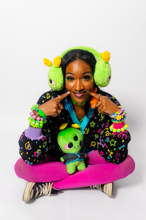 Girl sitting cross legged playfully looking at the camera. She is wearing bright green alien earmuffs, a roller rink carpet cardigan, pink tights and many chunky bracelets.