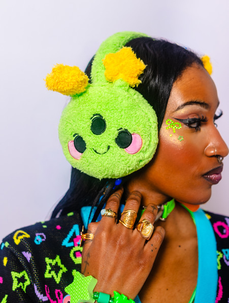 Girl from the shoulders up with her face to the side of the camera showing the alien plush ear muffs shes is wearing. In addition she has chunky green bracelets, a few rinks, and some matching green face gems.