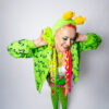 Girl with bright pink and green hair playfully looking down and away from the camera with her hands on her two low buns. She is wearing an alien green fuzzy headband with star antennae, a green bollo, a white tank top, a green slime cardigan, green and black striped tights, and black mary janes