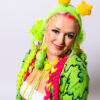 Girl from the waist up who has bright pink and green hair playfully looking down at the camera with her hands to her side. She is wearing an alien green fuzzy headband with star antennae, a green bollo, a white tank top, and a green slime cardigan.