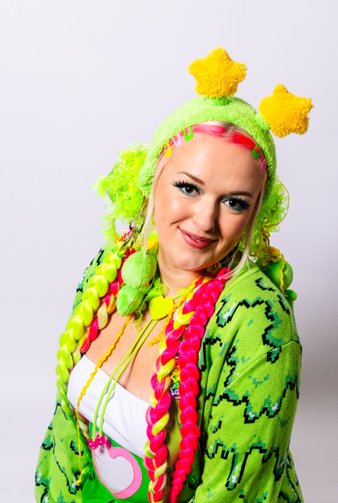 Girl from the waist up who has bright pink and green hair playfully looking down at the camera with her hands to her side. She is wearing an alien green fuzzy headband with star antennae, a green bollo, a white tank top, and a green slime cardigan.