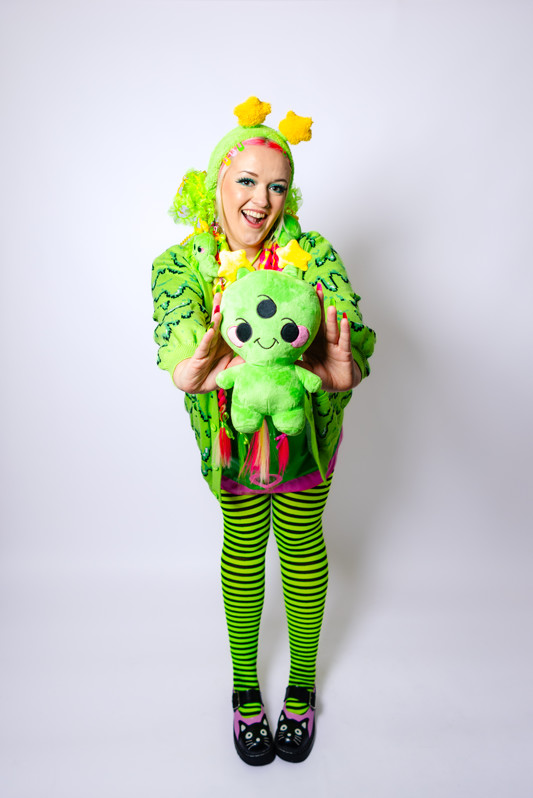 Girl playfully holding a cheerful alien plush toward the camera. She is wearing a matching alien green fuzzy headband with star antennae, a green slime cardigan, green and black striped tights and black mary janes