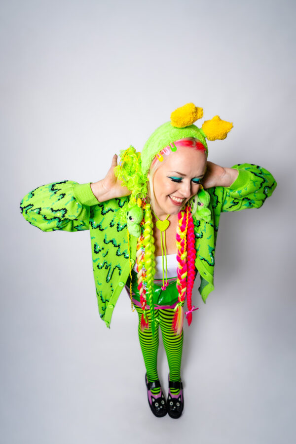 Girl with bright pink and green hair playfully looking down and away from the camera with her hands on her two low buns. She is wearing an alien green fuzzy headband with star antennae, a green bollo, a white tank top, a green slime cardigan, green and black striped tights, and black mary janes