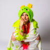 Woman with orange braids looking at the camera while crouching with one hand on her face. She is wearing an oversized cheerful alien plush hat with two star antennae, a green and white jacket, a green tank top, and white and green leg warmers.
