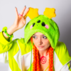 Woman from the chest up with orange braids looking slightly away from the camera with one hand touching the plush star on her hat. She is wearing an oversized cheerful alien plush hat with two star antennae, a green and white jacket and a green tank top.