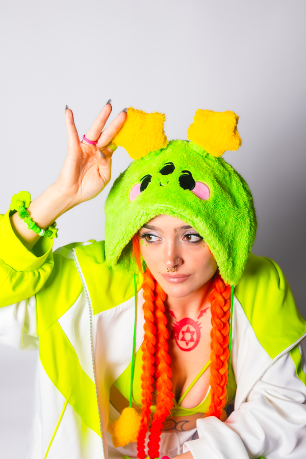 Woman from the chest up with orange braids looking slightly away from the camera with one hand touching the plush star on her hat. She is wearing an oversized cheerful alien plush hat with two star antennae, a green and white jacket and a green tank top.