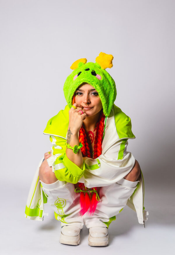 Woman with orange braids looking at the camera while crouching with one hand on her face. She is wearing an oversized cheerful alien plush hat with two star antennae, a green and white jacket, a green tank top, and white and green leg warmers.