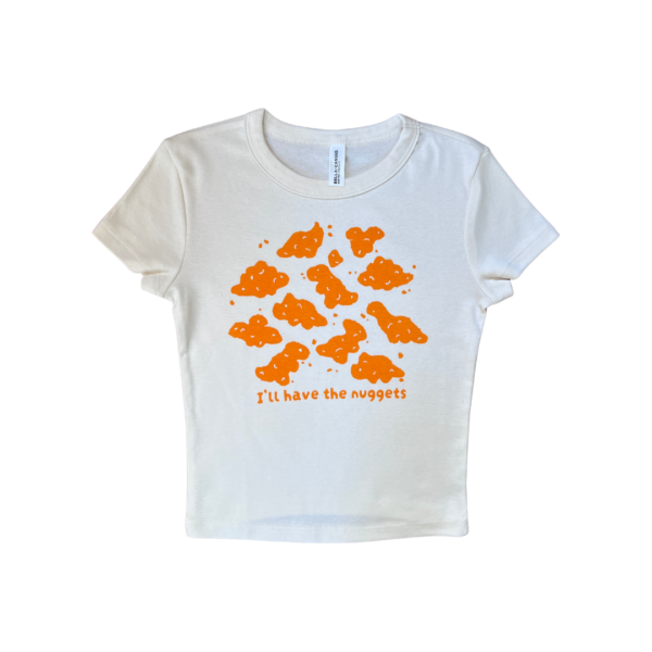 A cream-colored short-sleeve crop top with playful orange dino nuggets graphic and the text "I'll have the nuggets" on the front, laid flat on a clear background