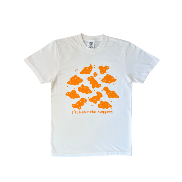 A cream-colored short-sleeve t-shirt with playful orange dino nuggets graphic and the text "I'll have the nuggets" on the front, laid flat on a clear background.