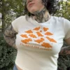 Woman wearing a cream-colored short-sleeve baby tee with playful orange dinosaur nuggets graphic and the text "I'll have the nuggets" and a jean skirt outside with grass in the background.