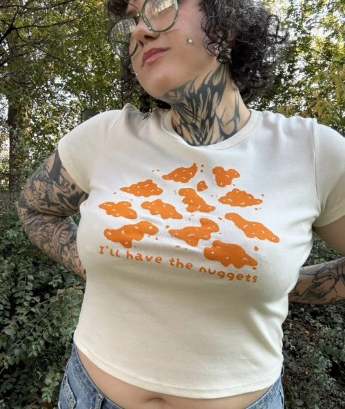 Woman wearing a cream-colored short-sleeve baby tee with playful orange dinosaur nuggets graphic and the text "I'll have the nuggets" and a jean skirt outside with grass in the background.