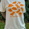 Woman wearing a cream-colored short-sleeve t-shirt with playful orange dinosaur nuggets graphic and the text "I'll have the nuggets" and a jean skirt outside with grass in the background.
