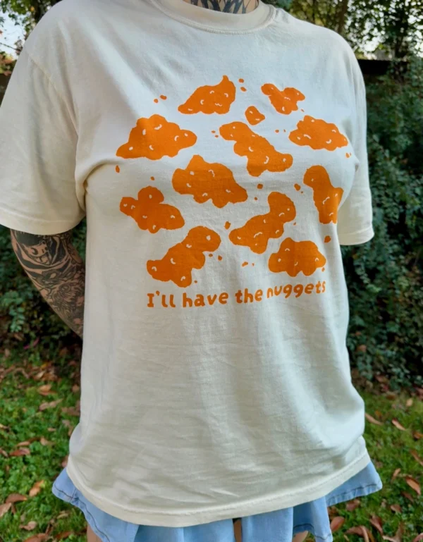 Woman wearing a cream-colored short-sleeve t-shirt with playful orange dinosaur nuggets graphic and the text "I'll have the nuggets" and a jean skirt outside with grass in the background.