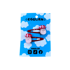 A pair of playful mushroom cabochons on red hairclips both on a blue backing card that reads "Egglien"
