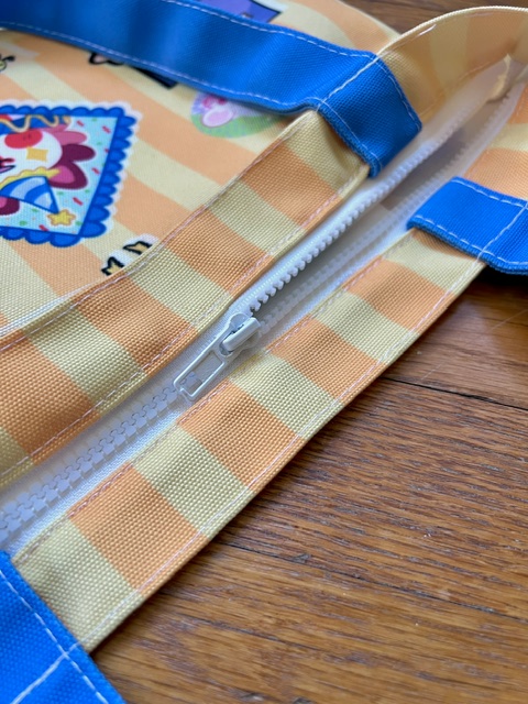 A close up view of a yellow striped tote bag with blue straps laying on wood floor. The bag has a white zipper that is half open.