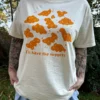 Woman wearing a cream-colored short-sleeve t-shirt with playful orange dinosaur nuggets graphic and the text "I'll have the nuggets" and a jean skirt outside with grass in the background.