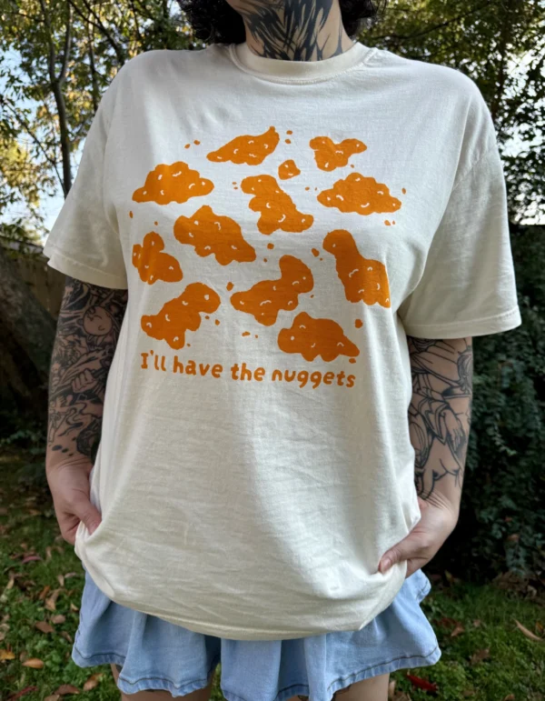 Woman wearing a cream-colored short-sleeve t-shirt with playful orange dinosaur nuggets graphic and the text "I'll have the nuggets" and a jean skirt outside with grass in the background.